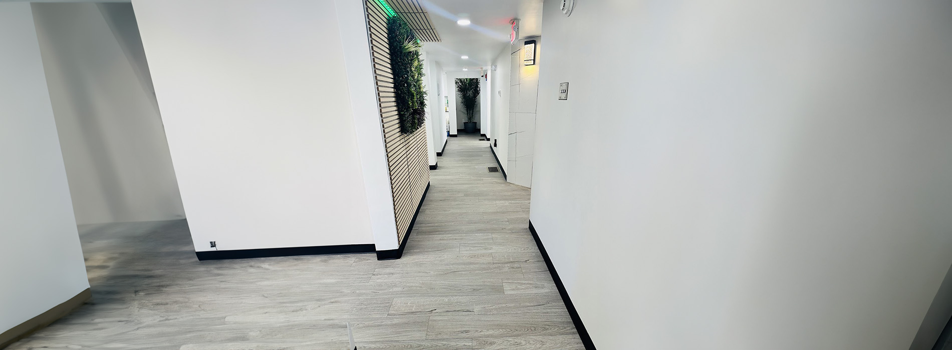 The image shows a modern interior hallway with a blurred view of a white wall, a wooden floor, and a long corridor leading to an open door at the end.