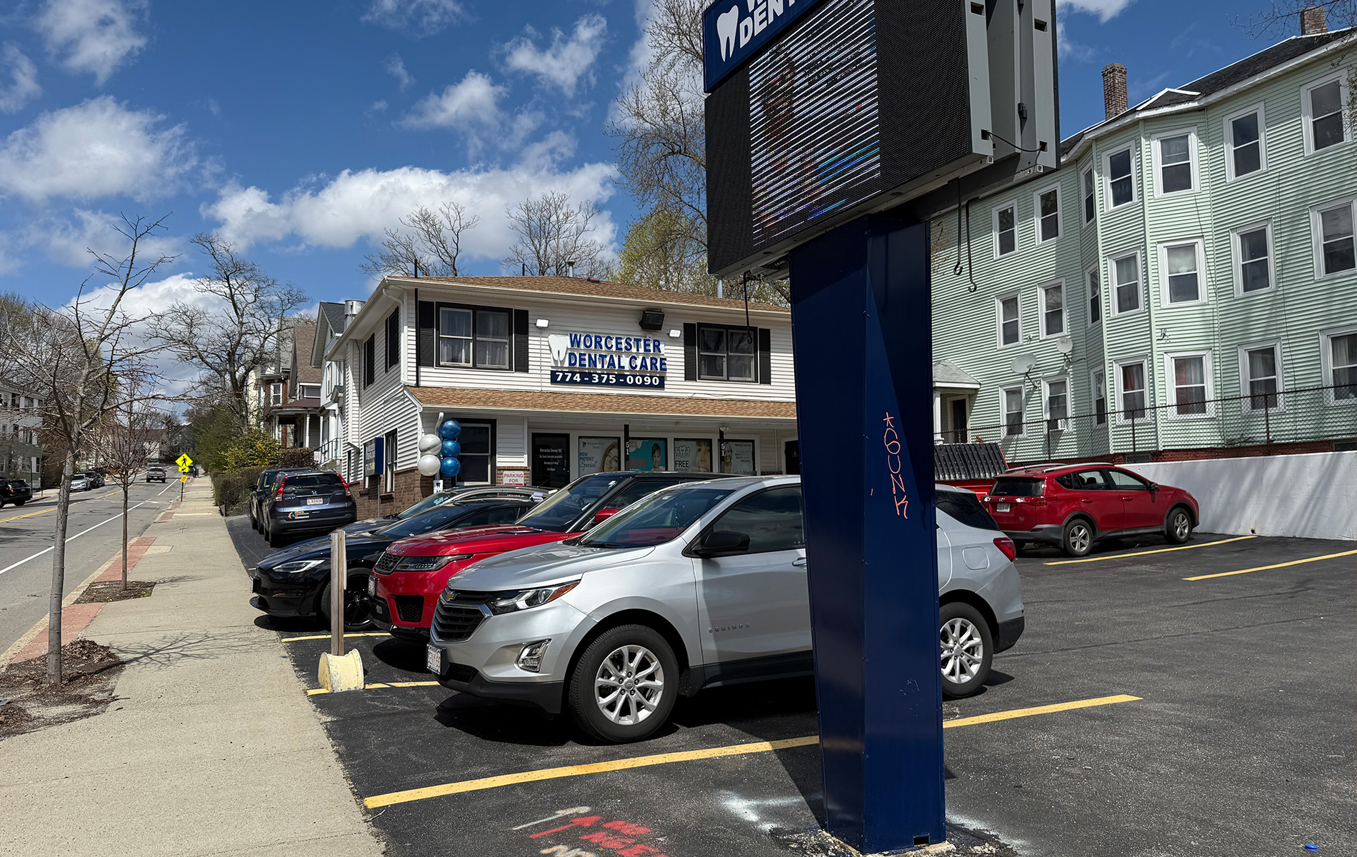 The image shows a parking lot with several cars parked and a large electronic scoreboard displaying a game s score, located next to a street corner with a storefront and a sign that reads  WINGS .