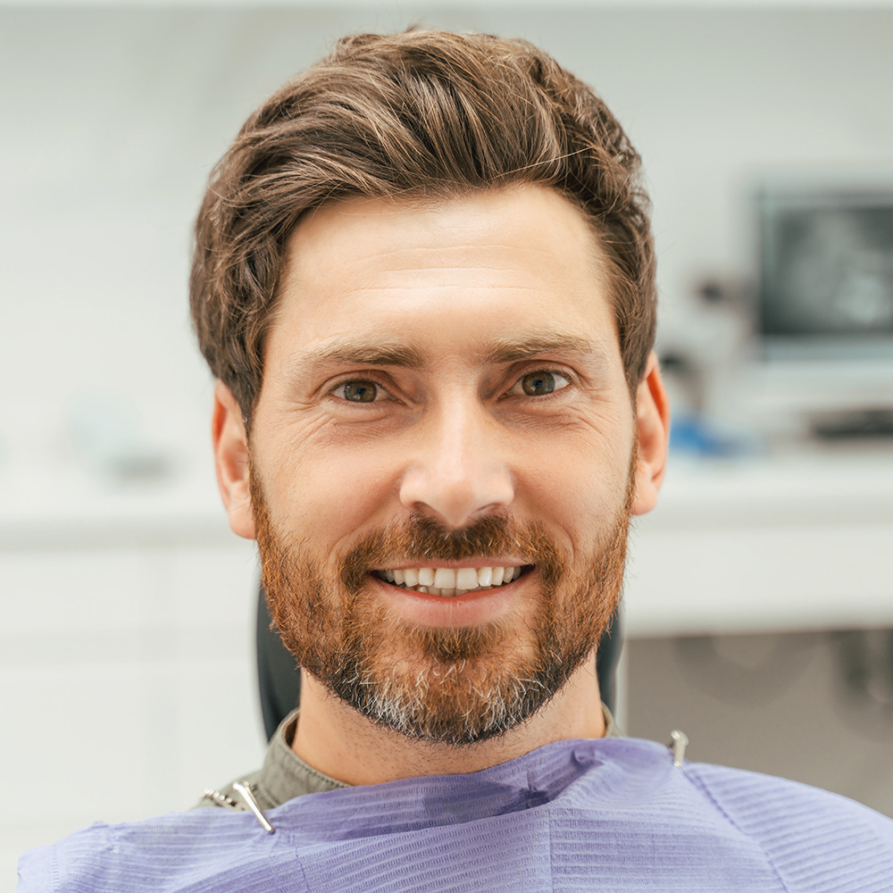 The image shows a man with short brown hair and a beard, wearing glasses and sitting in a dental chair, smiling at the camera.
