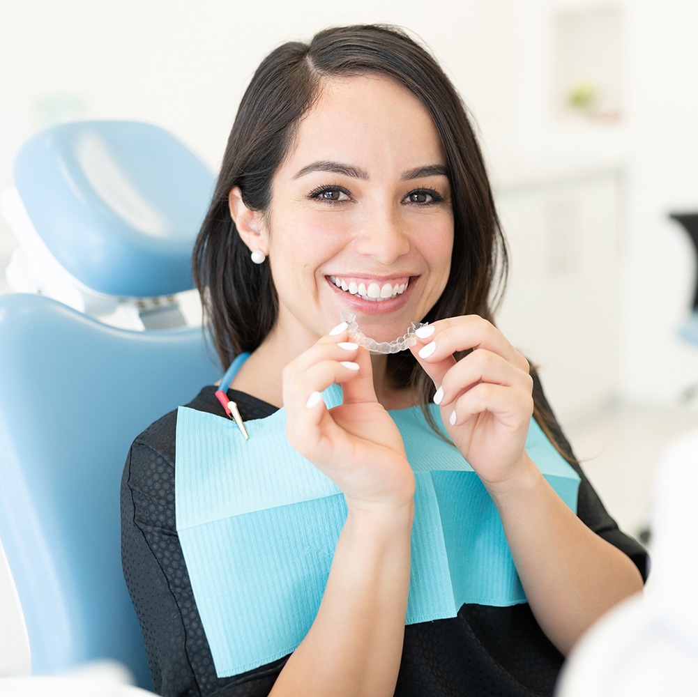 This is an image of a woman sitting in a dental chair, holding a toothbrush with toothpaste on her teeth, smiling at the camera while wearing a blue face mask.