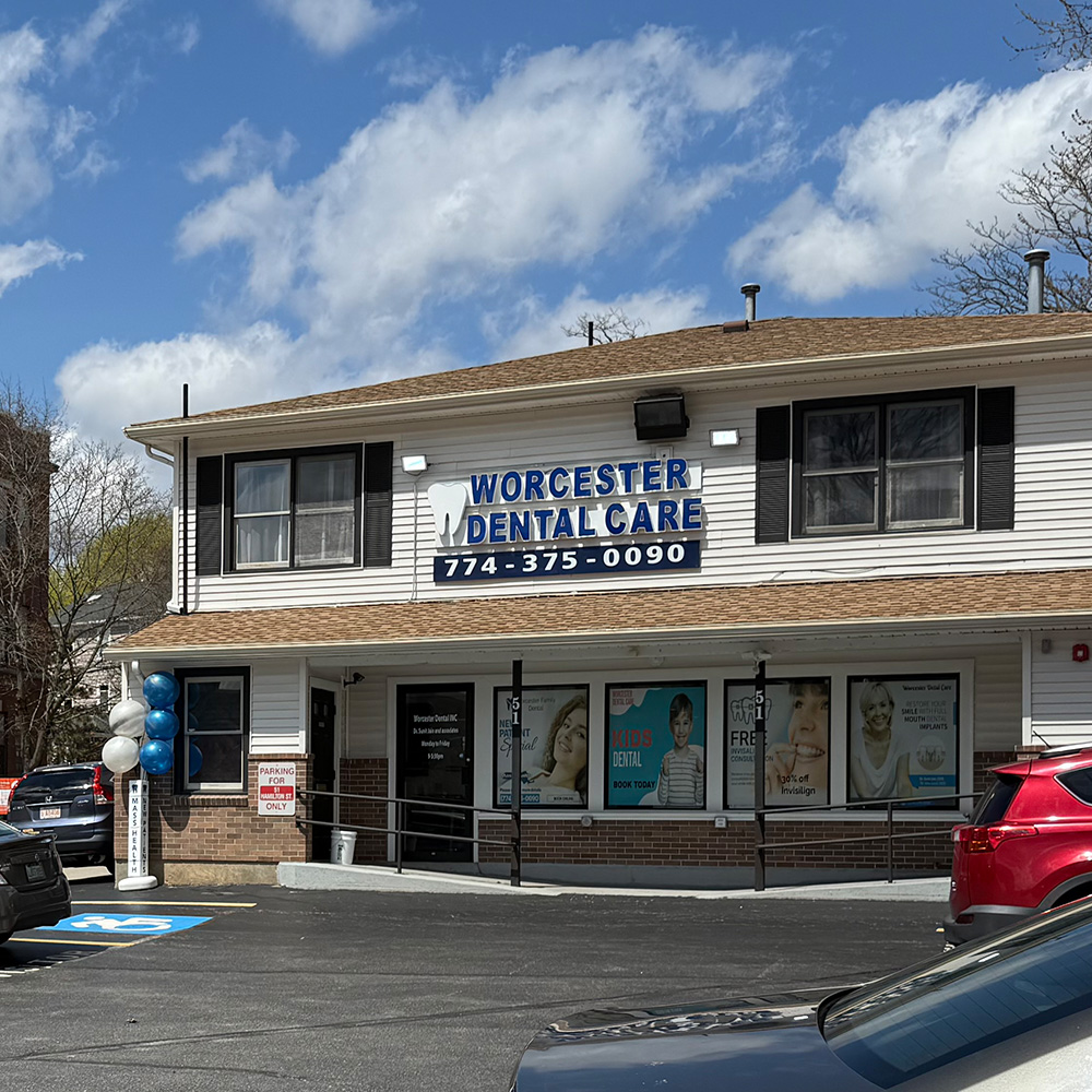 The image shows a dental practice named 'Worcester Dental Care' with a sign indicating its location at 103 Main Street, Worcester, Massachusetts. The building has a parking lot with cars parked in front of it and is situated under a cloudy sky.