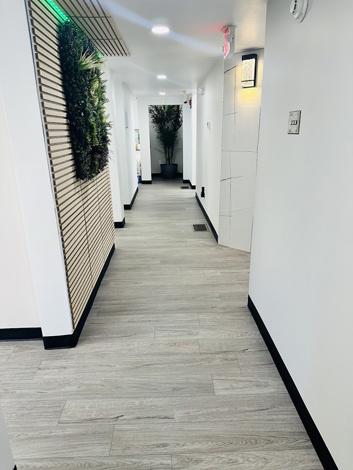 The image shows an interior hallway with a long, rectangular floor tile pattern, white walls, and a corridor leading to a door at the end.