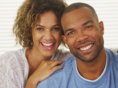 The image shows a man and a woman smiling at each other  the man has a beard and is wearing a light blue shirt, while the woman has curly hair and is dressed in a white top with lace detailing.